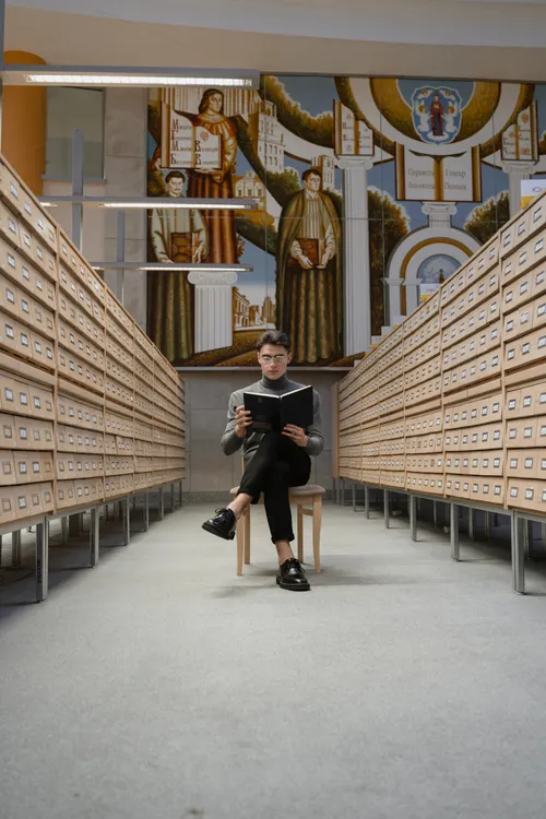 A student reading in a grand library between card catalog shelves and a mural
